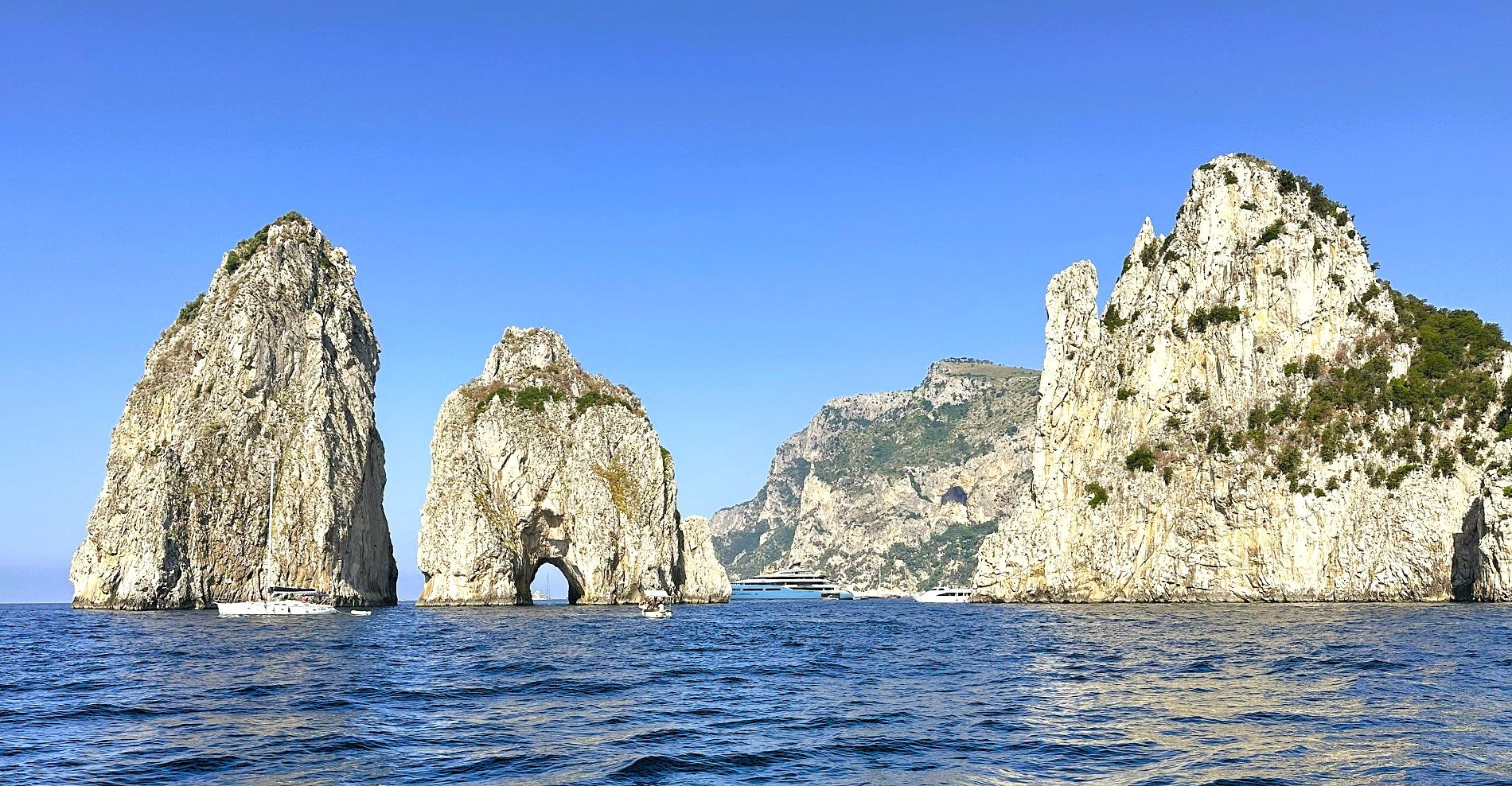 View of the Faraglioni sea stacks off the coast of Capri, Italy, with boats and rugged cliffs on a clear Mediterranean day.
