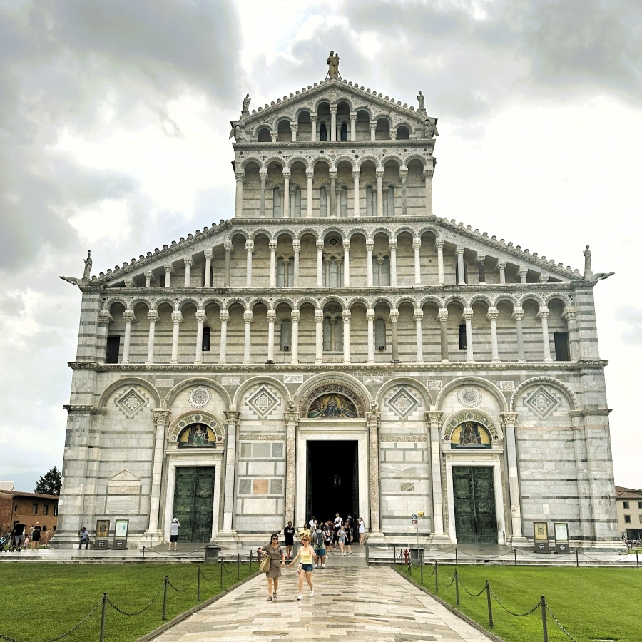 Front view of Pisa Cathedral in Piazza dei Miracoli showing its Romanesque marble façade with three bronze doors and rows of arches under cloudy sky