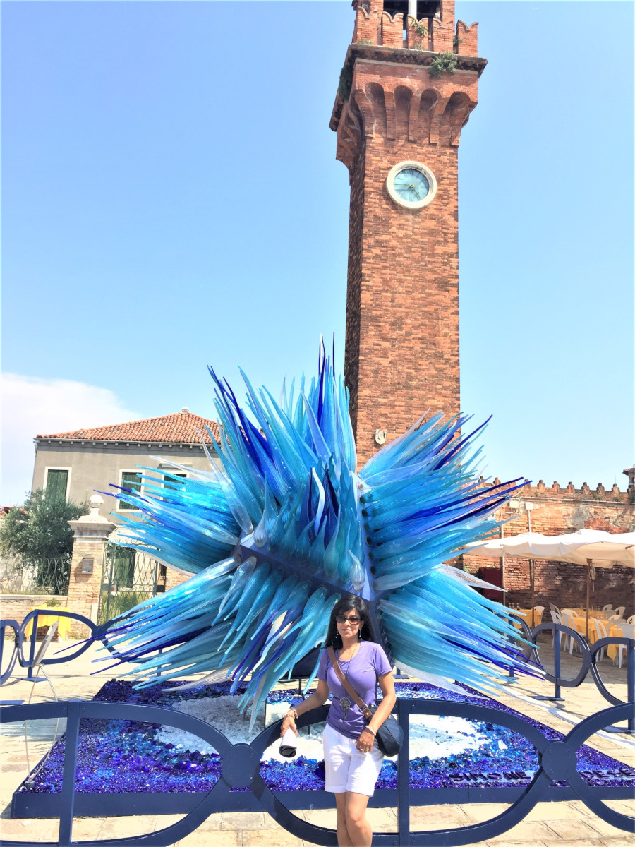 Traveler standing near the Murano Clock Tower beside the striking blue glass sculpture in Campo Santo Stefano, a landmark of Venetian glass art