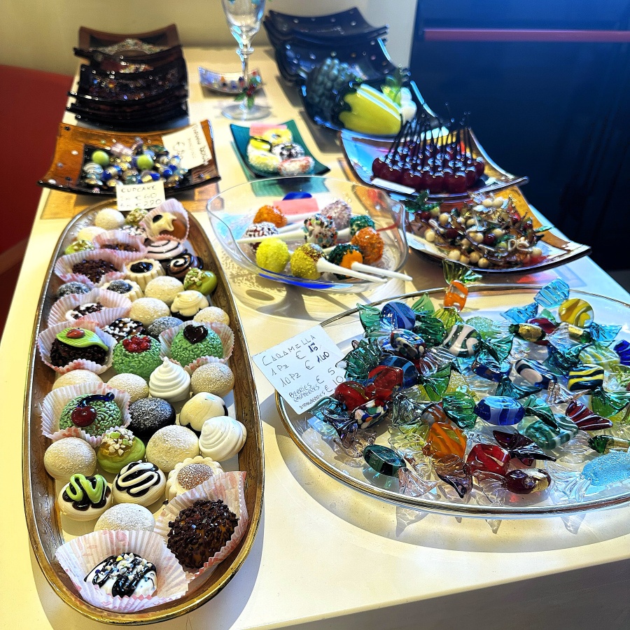 Assorted Murano glass candies displayed alongside colorful confectionery in a souvenir shop on Murano Island, Italy.