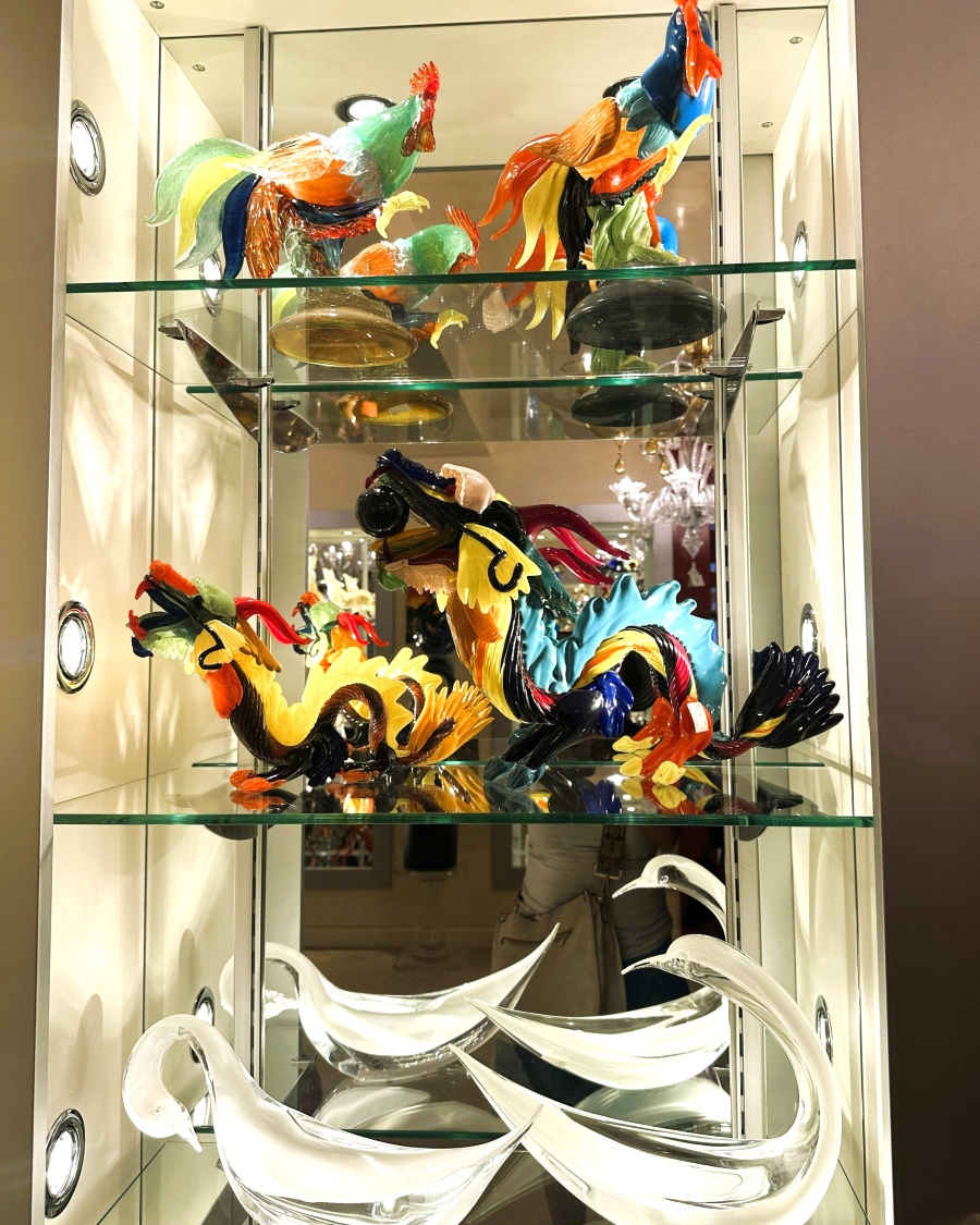 Colorful Murano glass roosters and clear swans displayed in a glass cabinet, showcasing the artistry and precision of Venetian glassmakers.