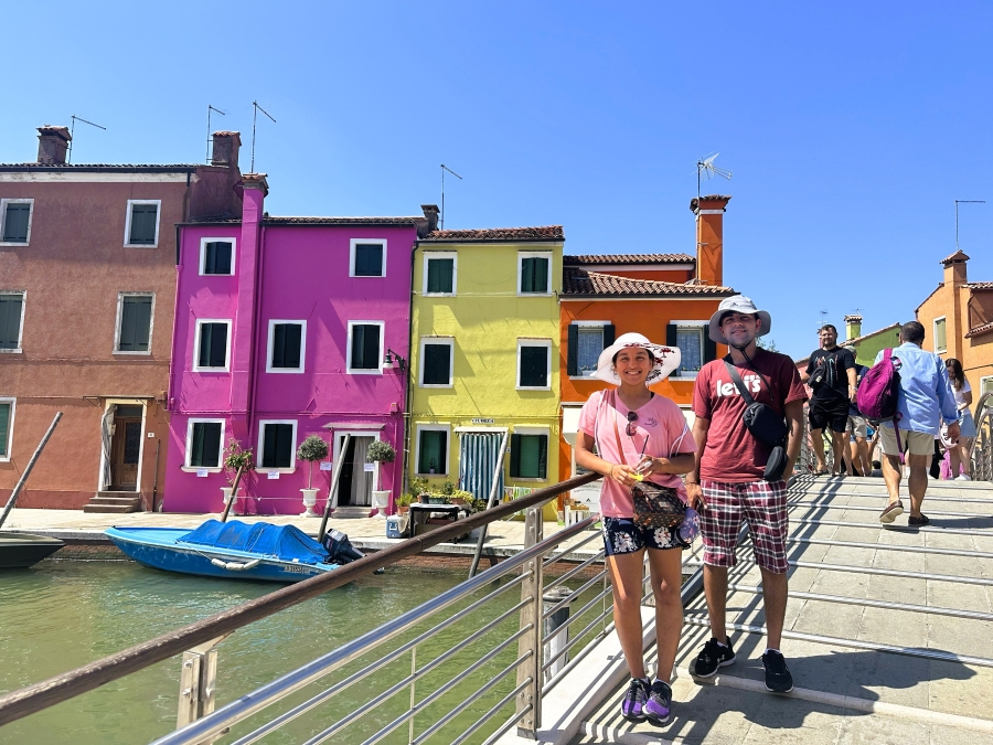 Brightly painted houses along Burano’s canal with travelers standing on a bridge under the Venetian sun