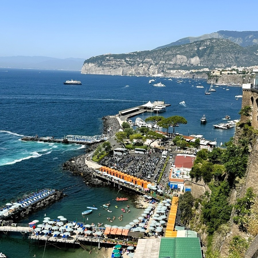 A panoramic view of Sorrento’s Marina Grande with cliffs, beach clubs, and blue Tyrrhenian Sea waters on a sunny summer day.