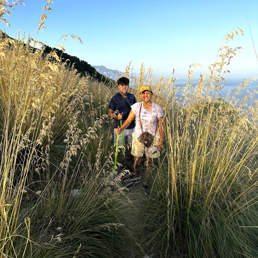 Hikers walking through tall golden Mediterranean grasses along the Sorrento Peninsula sunset trail with sea views in the background.