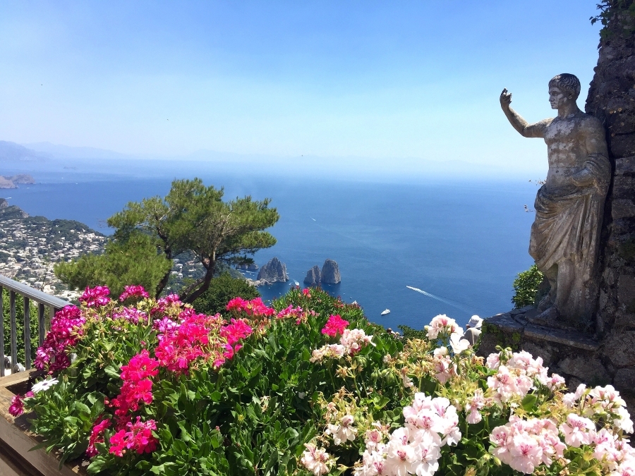 View from Monte Solaro in Anacapri featuring the iconic statue overlooking the Faraglioni sea stacks and vibrant flowers in the foreground.